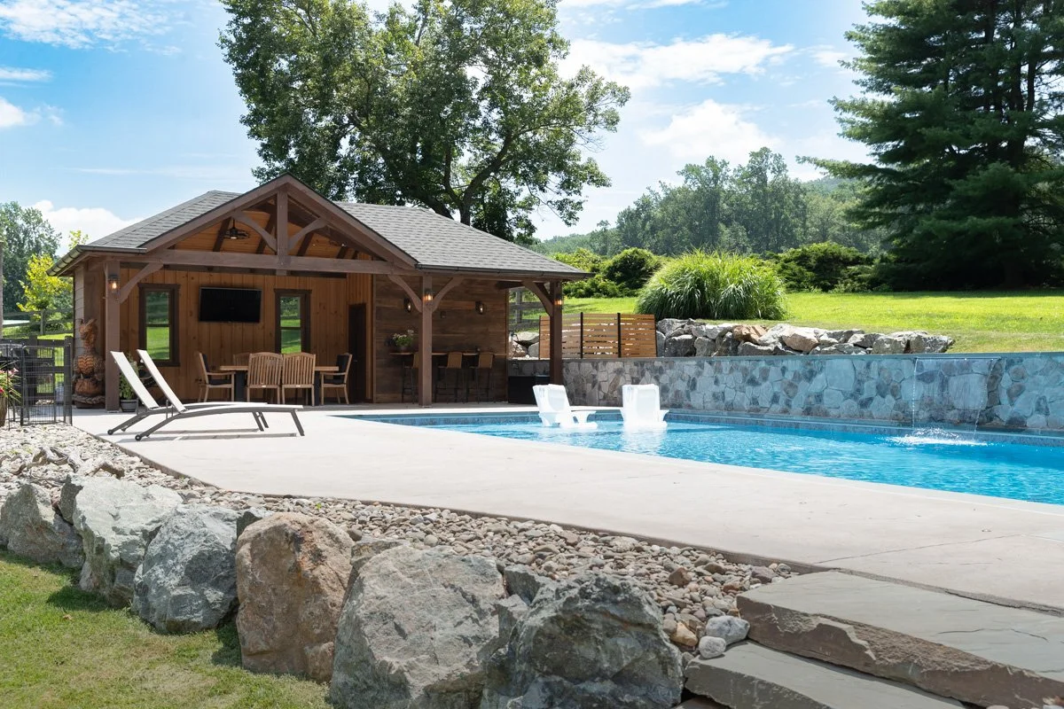 A backyard swimming pool with a wooden pool house, lounge chairs, and surrounding rocks and greenery.