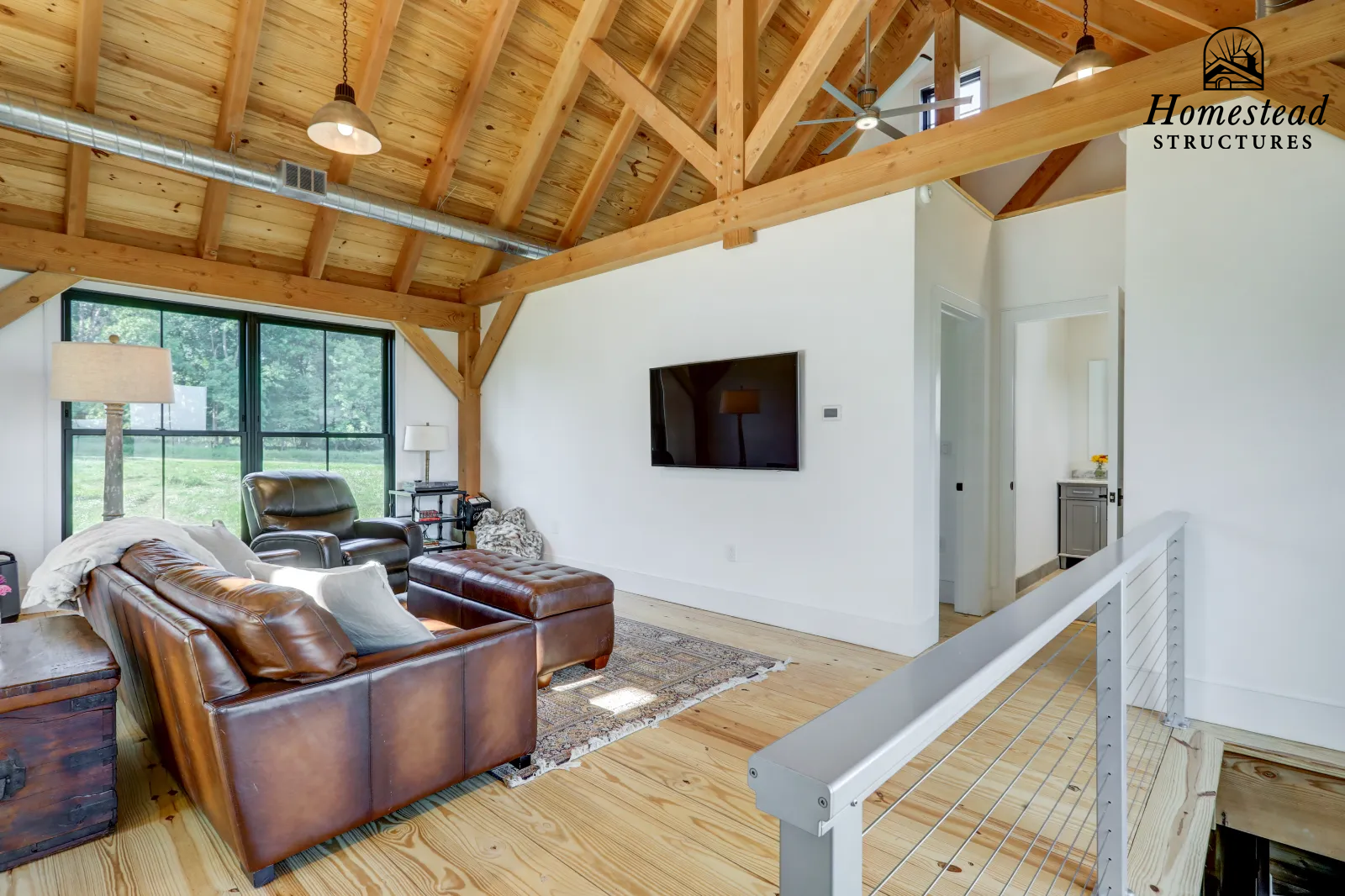 Living room with wooden ceiling beams and a large window showing green outdoor scenery. Contains a leather sofa, a black armchair, a side table with a lamp, and a wall-mounted TV.