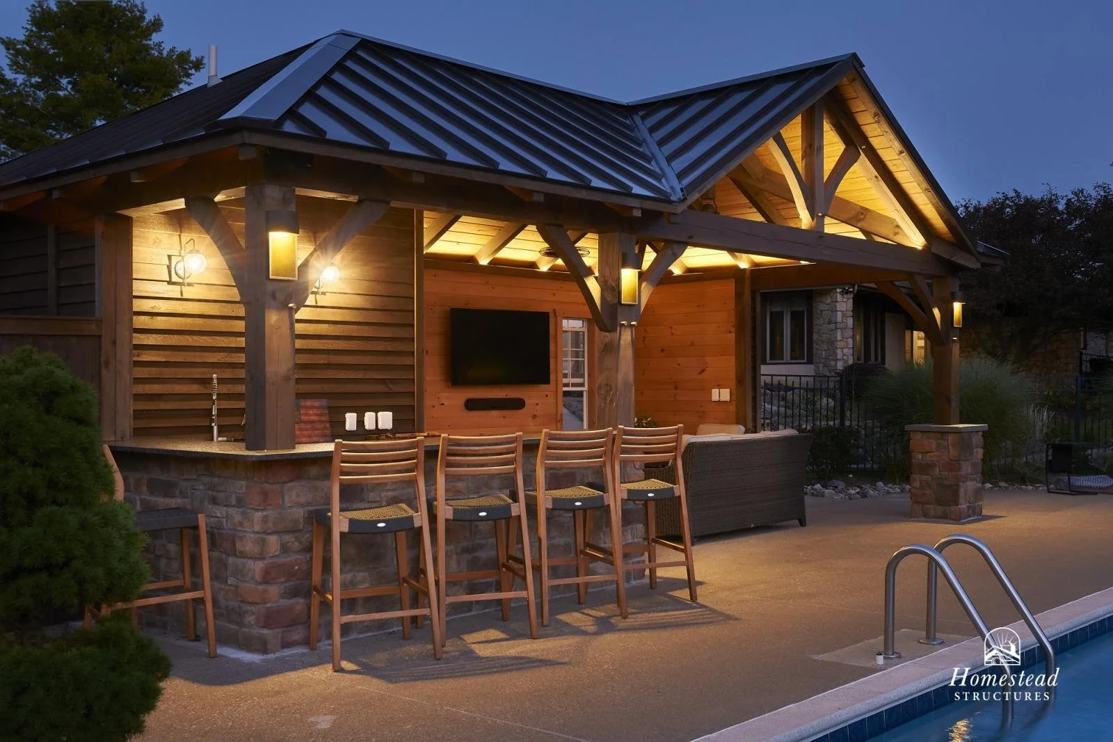 Outdoor poolside area at dusk featuring a wooden pergola with lighting, bar seating, a wall-mounted TV, and seating area, with a swimming pool in the foreground.