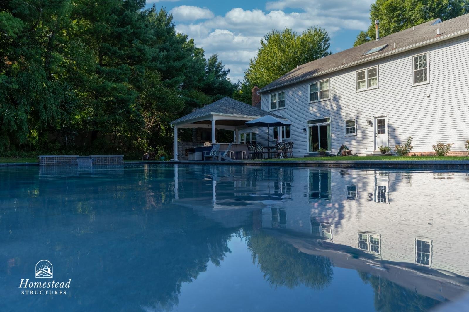 A backyard swimming pool reflecting a white two-story house with multiple windows and a sliding glass door, surrounded by green trees and a wooden deck with outdoor furniture and an umbrella.