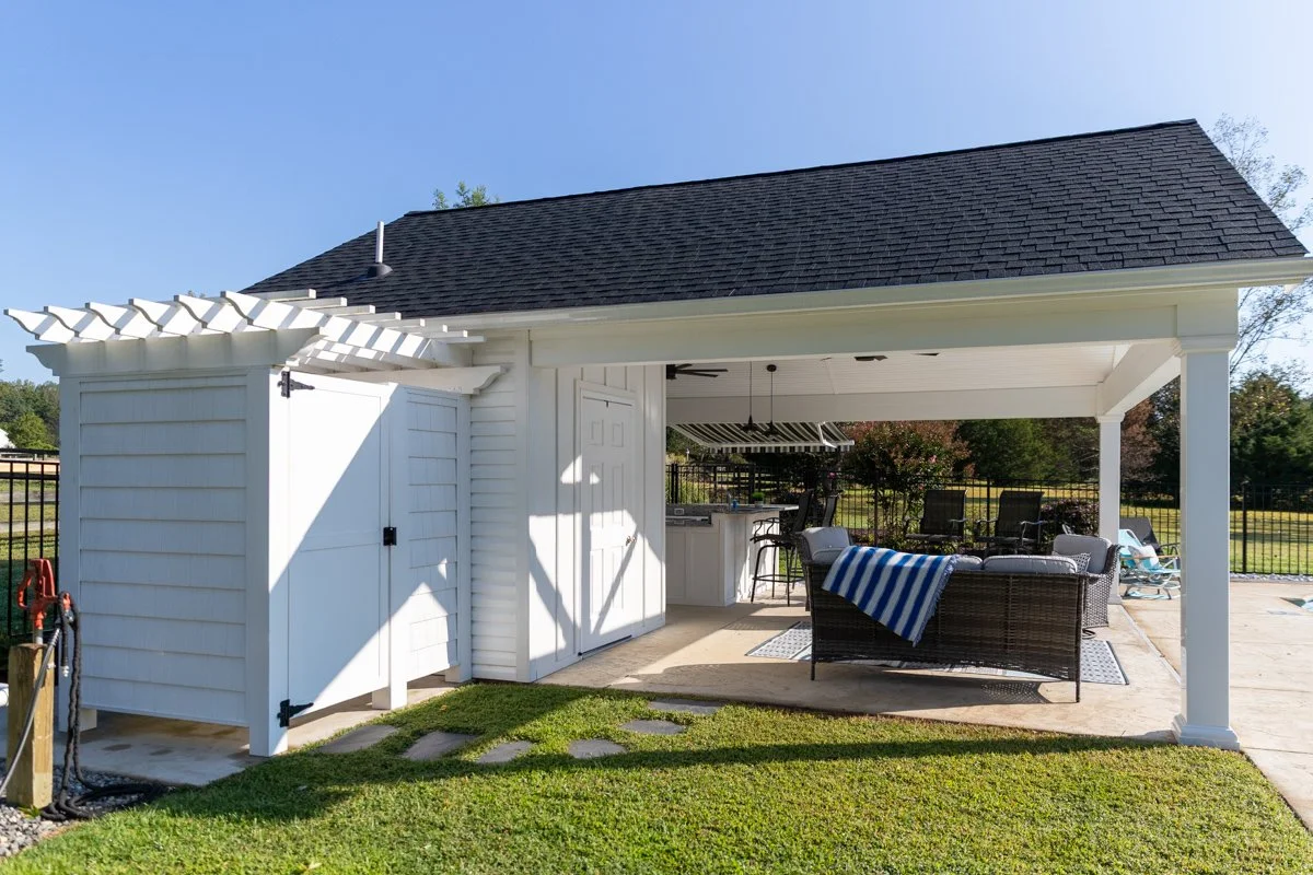 Outdoor patio with white pavilion, wicker couch with striped blanket, chairs, and a built-in kitchen area with barbecue grill, surrounded by a black metal fence and green lawn.