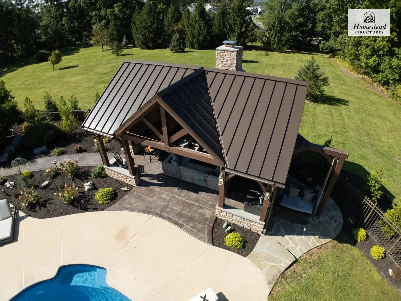 A backyard patio with a metal-roofed pavilion, outdoor furniture, and a swimming pool, surrounded by landscaped garden beds and green lawn, with trees and a stone chimney in the background.