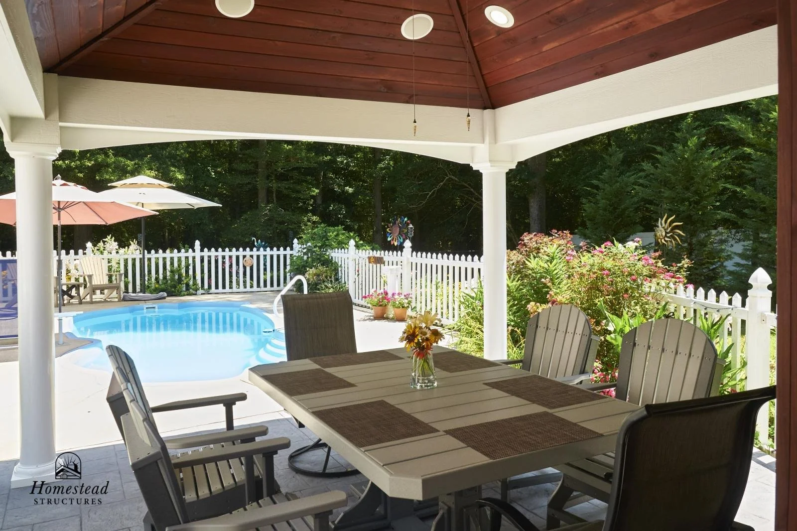 Covered patio with outdoor dining table, surrounded by a backyard with a swimming pool, pink and purple flowers, and a white picket fence with trees beyond.