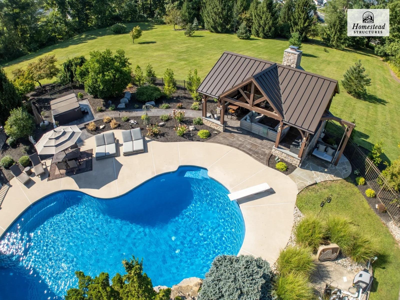 Aerial view of a backyard with a kidney-shaped swimming pool, poolside lounge chairs, an umbrella, a hot tub, a covered patio area with seating, and a large grassy yard with trees.