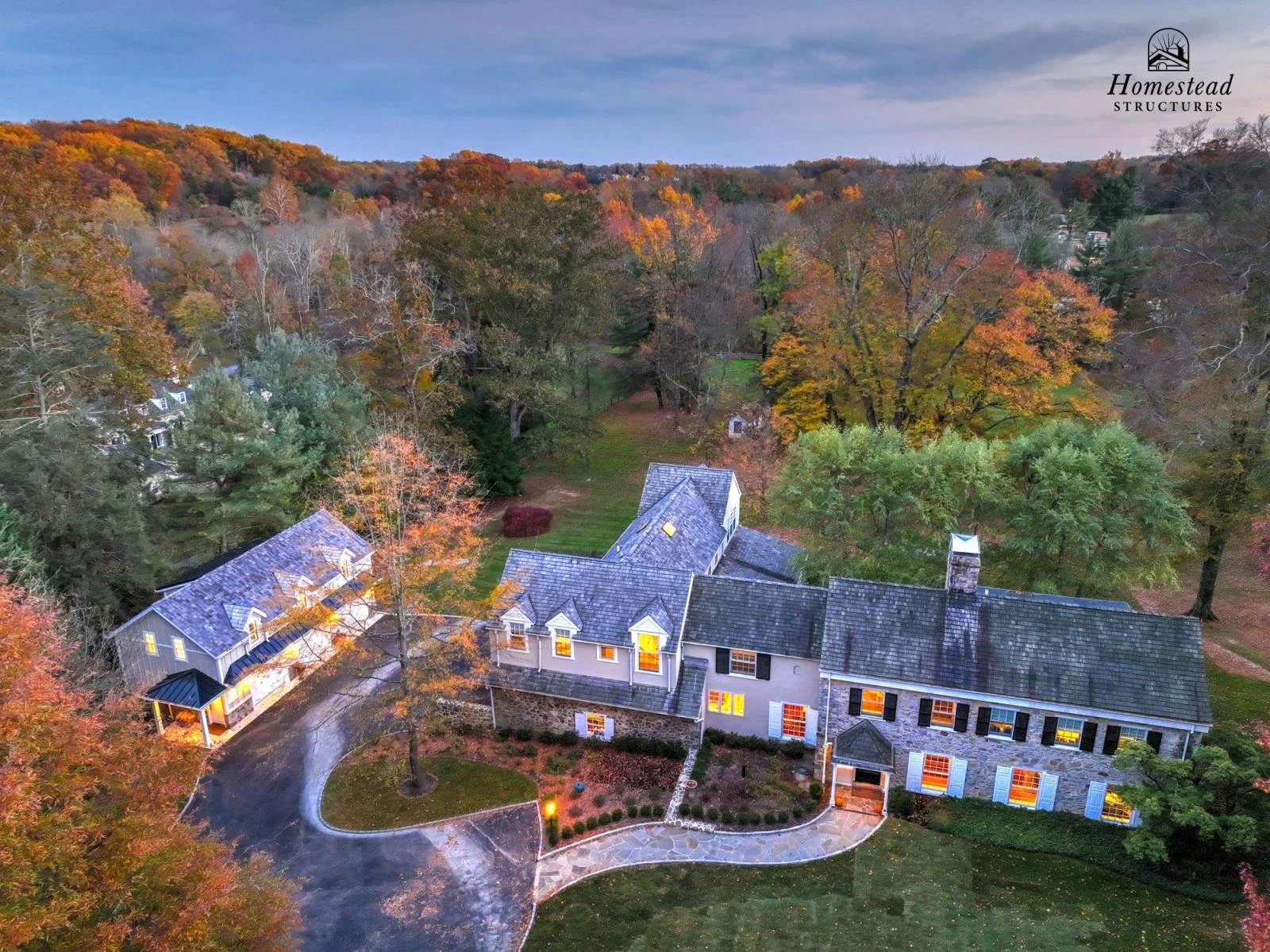A large house with a stone and siding exterior, multiple gabled roofs, and lit windows, surrounded by trees with fall foliage, a curved driveway, and a lush green lawn, set in a wooded area during dusk.