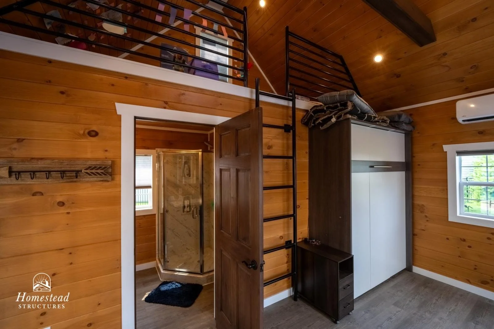 Interior view of a wooden cabin room with a white wardrobe, loft area accessible by a ladder, and a bathroom with a glass shower stall, a window, and an air conditioning unit on the wall.