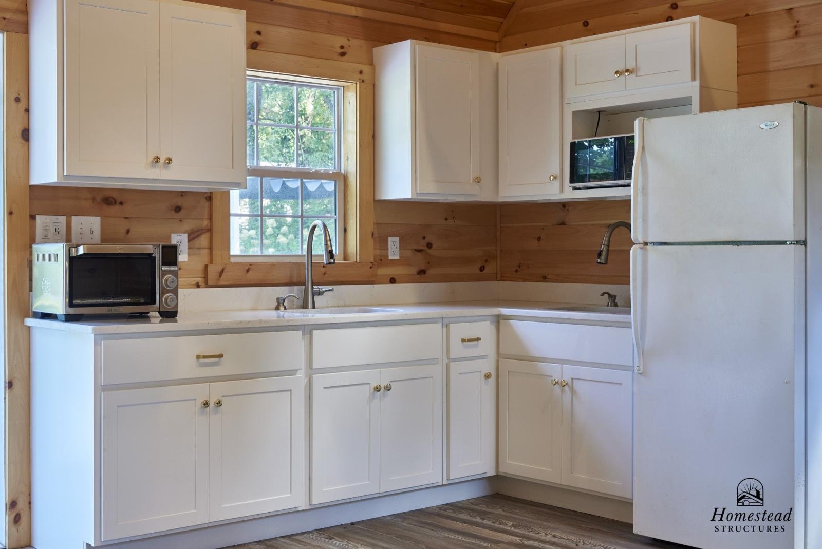 Kitchen with white cabinets, a small microwave, a refrigerator, a window showing greenery outside, and wood-paneled walls.
