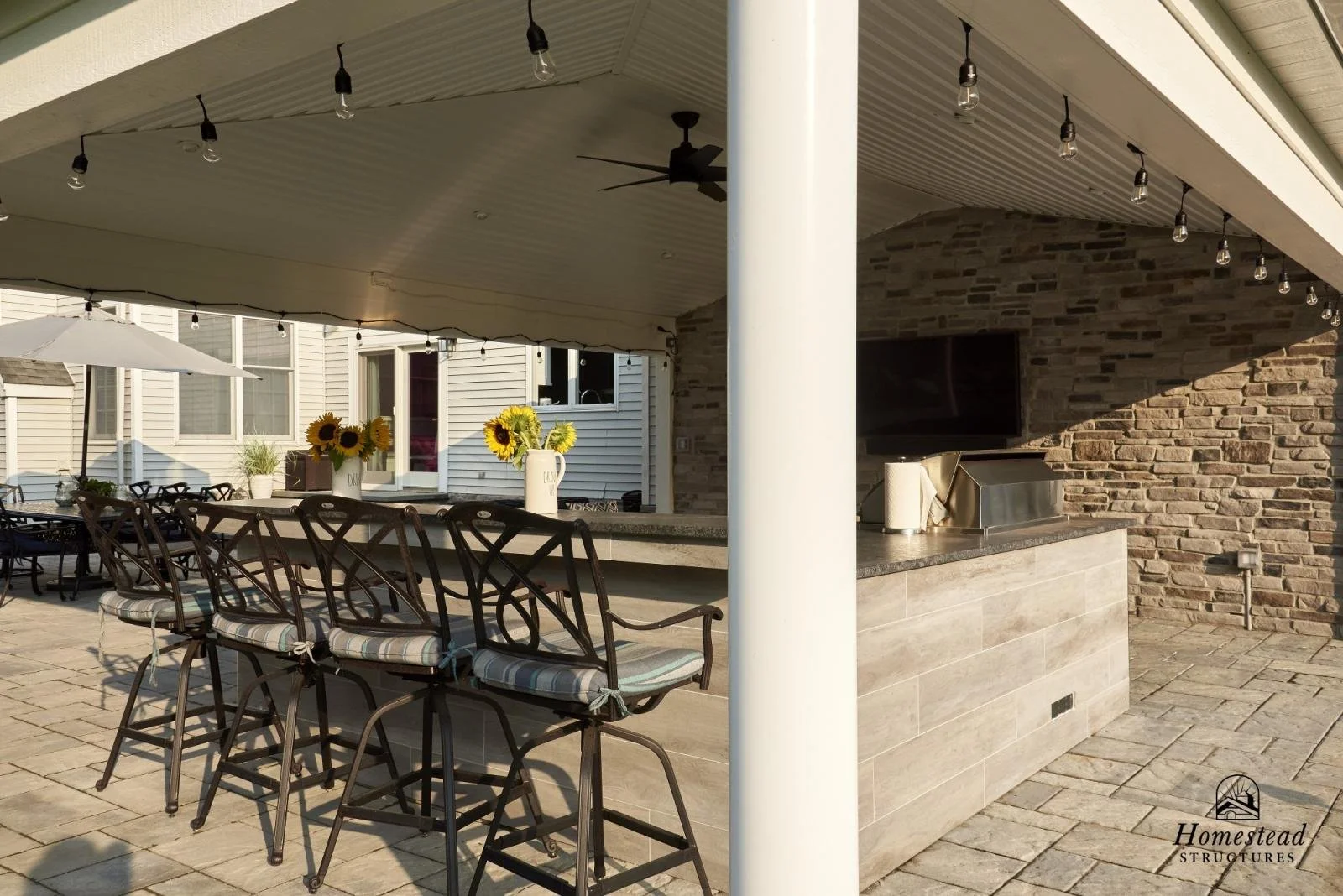An outdoor patio area with bar seating, sunflowers in vases, string lights, a ceiling fan, an umbrella, and a brick wall with a mounted TV.