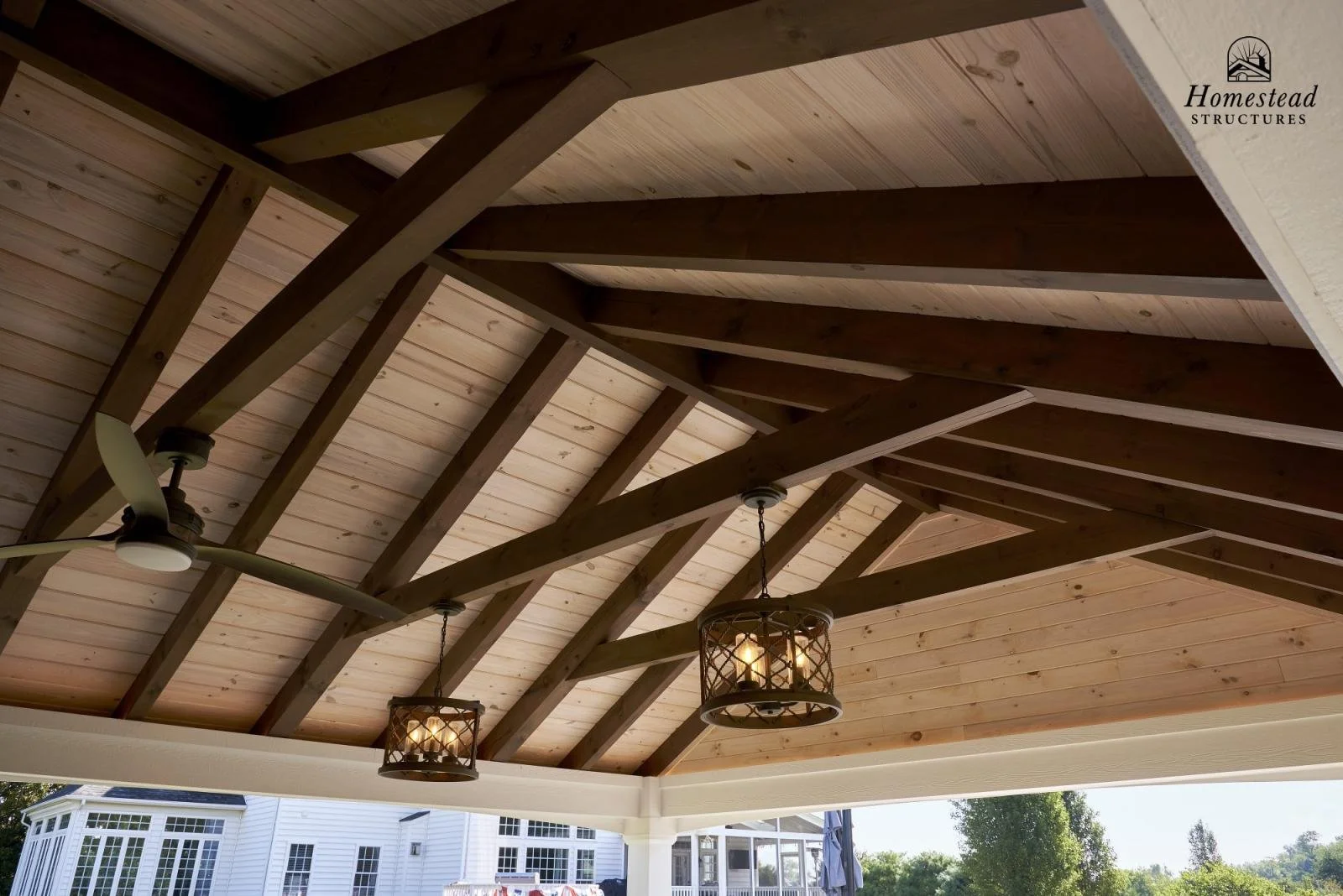 View of a wooden ceiling with exposed beams, ceiling fans, and hanging light fixtures under a porch or patio, with outdoor scenery visible in the background.