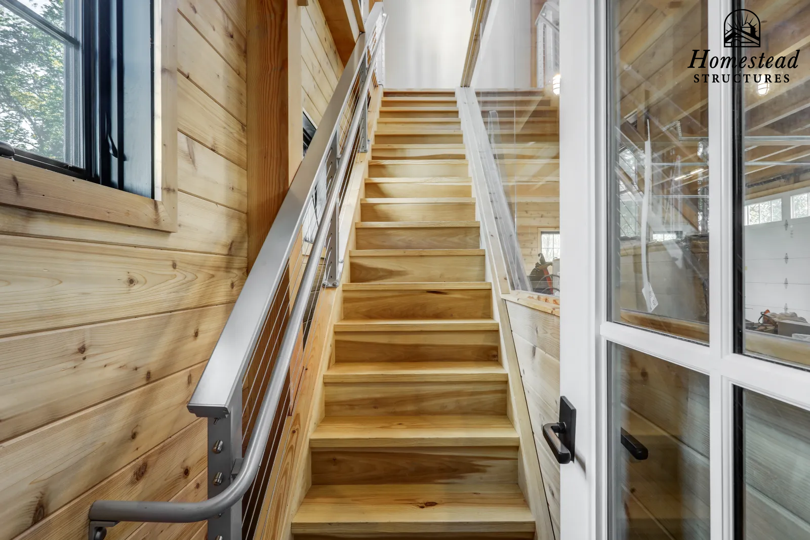 Interior view of a wooden staircase with metal handrails leading up. The walls are paneled with light wood, and there are windows on the left and right sides. The logo of Homestead Structures is in the upper right corner.