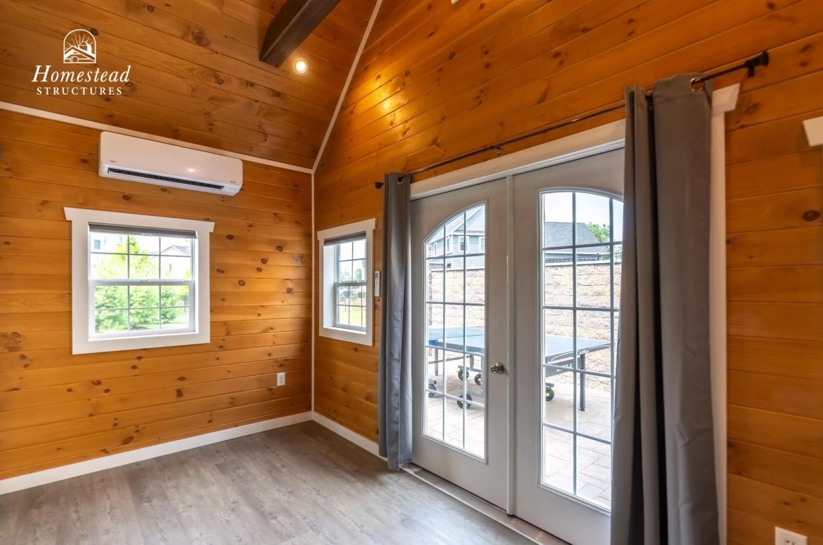 Interior of a room with wooden paneled walls, a ceiling-mounted air conditioner, two small windows with white trims, and large glass door with grid pattern, gray curtains, and a view of an outdoor patio with a ping pong table.