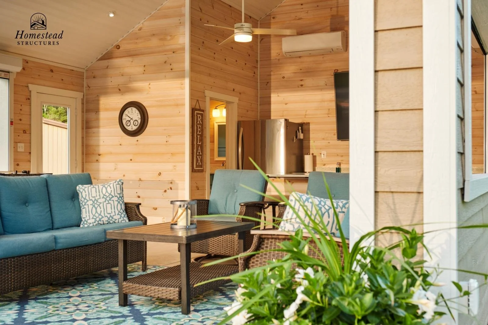 Living room with wooden walls, blue cushioned sofas, patterned pillows, a wooden coffee table, and potted plant in the foreground. Kitchen with stainless steel refrigerator and a wall-mounted TV visible in the background.
