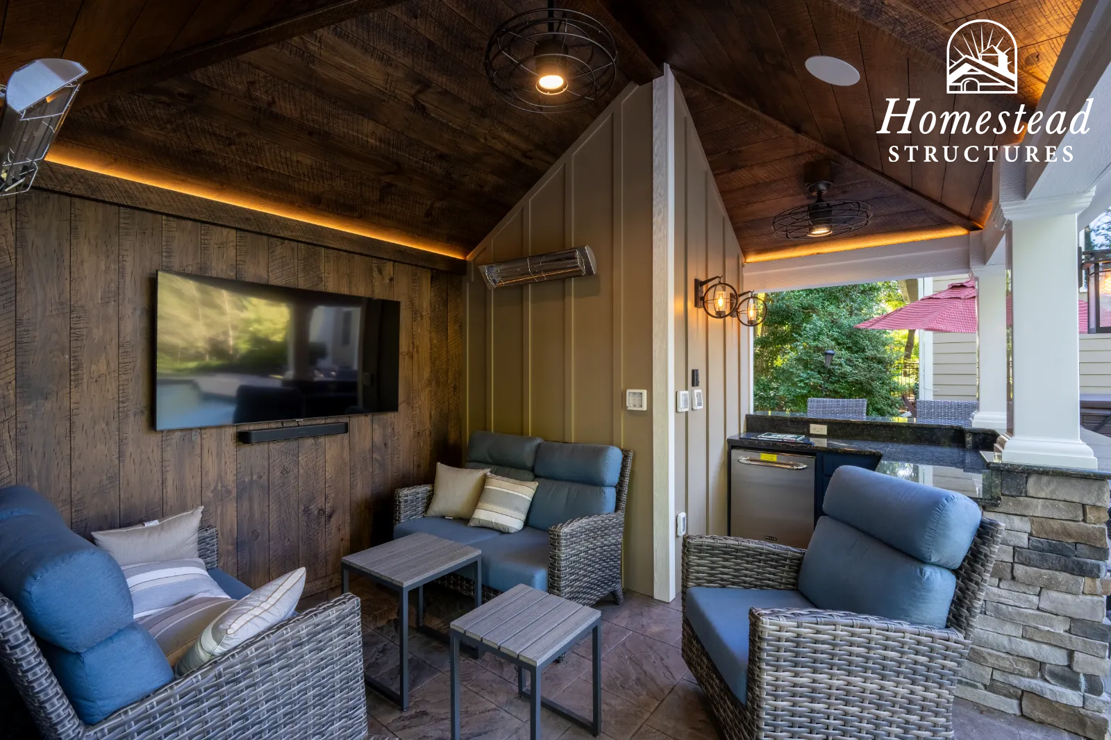 Outdoor living space with wicker armchairs, a sofa with pillows, a wall-mounted television, and a small outdoor kitchenette with a mini fridge, under a wooden ceiling with built-in lighting and a ceiling fan.