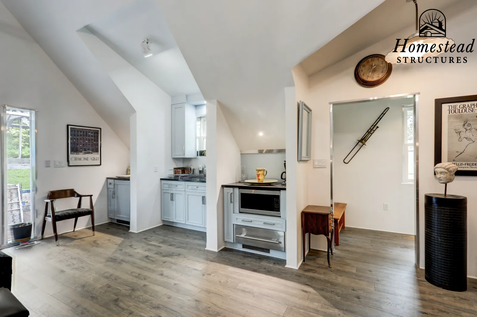 Interior view of a home with white walls, light wooden flooring, a small kitchen area with white cabinets, a microwave, a clock on the wall, art pieces, and decorative objects.