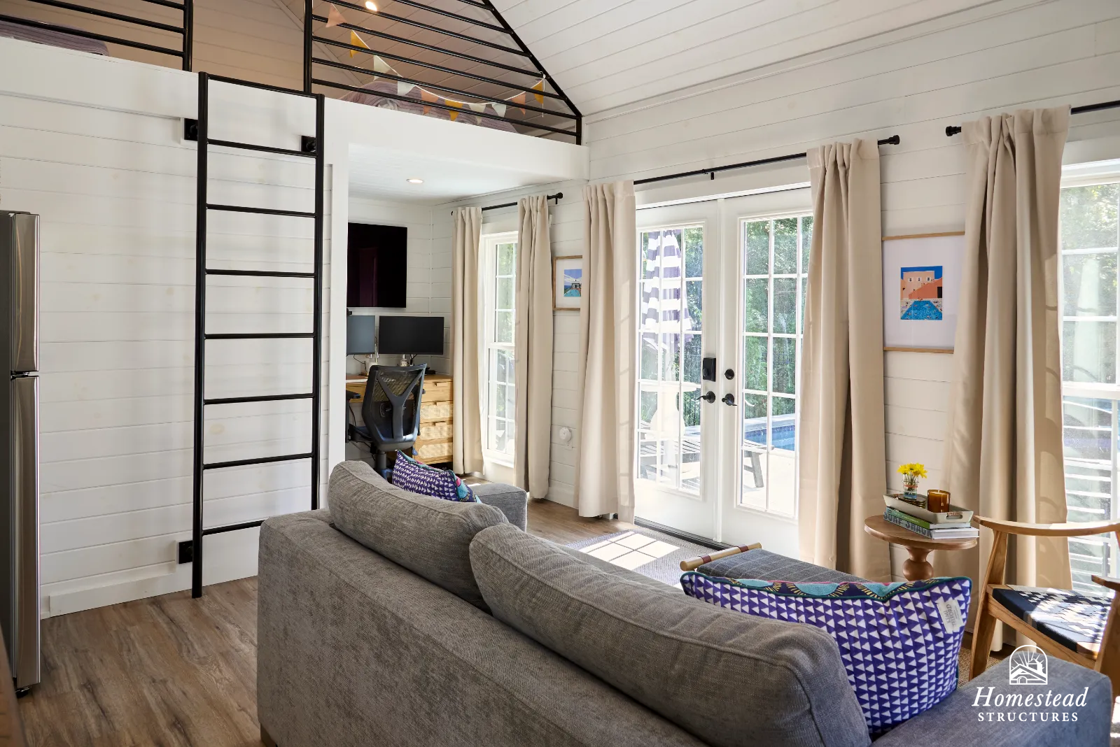 Living room with glass door leading to outdoor space, cream curtains, a gray couch with patterned pillows, and a small side table with books and a vase with flowers.
