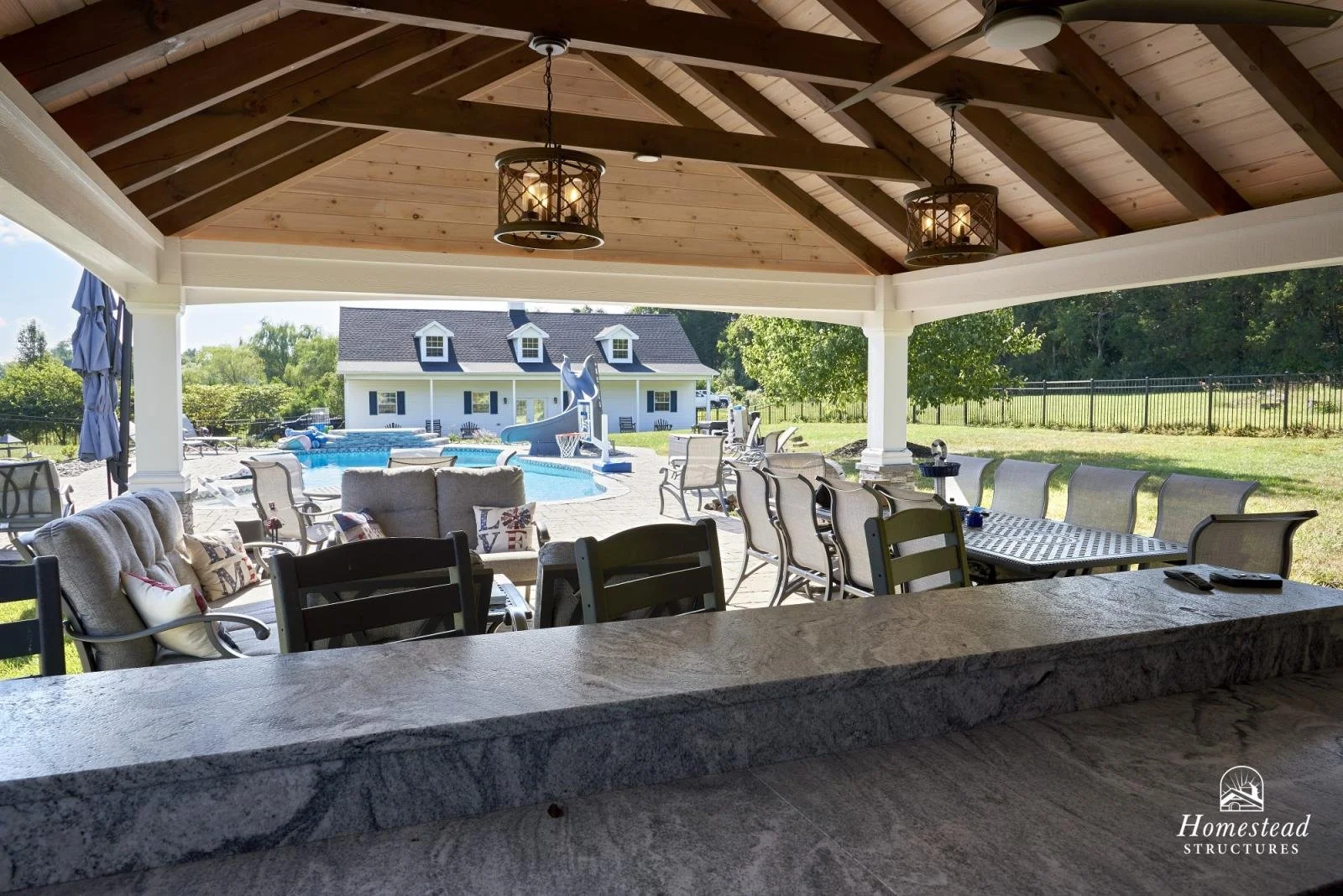 Covered outdoor patio with a granite countertop, ceiling fans, and a view of a backyard pool, gazebo, and white house.