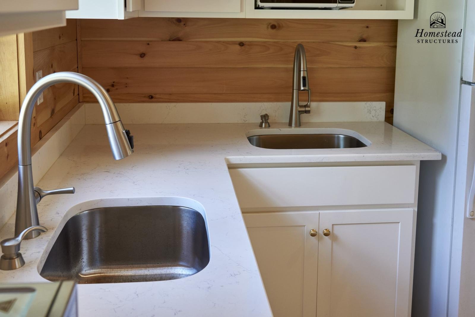 Kitchen countertop with two stainless steel sinks, wooden panel wall, white cabinets, and a refrigerator on the right. The logo of Homestead Structures is in the upper right corner.