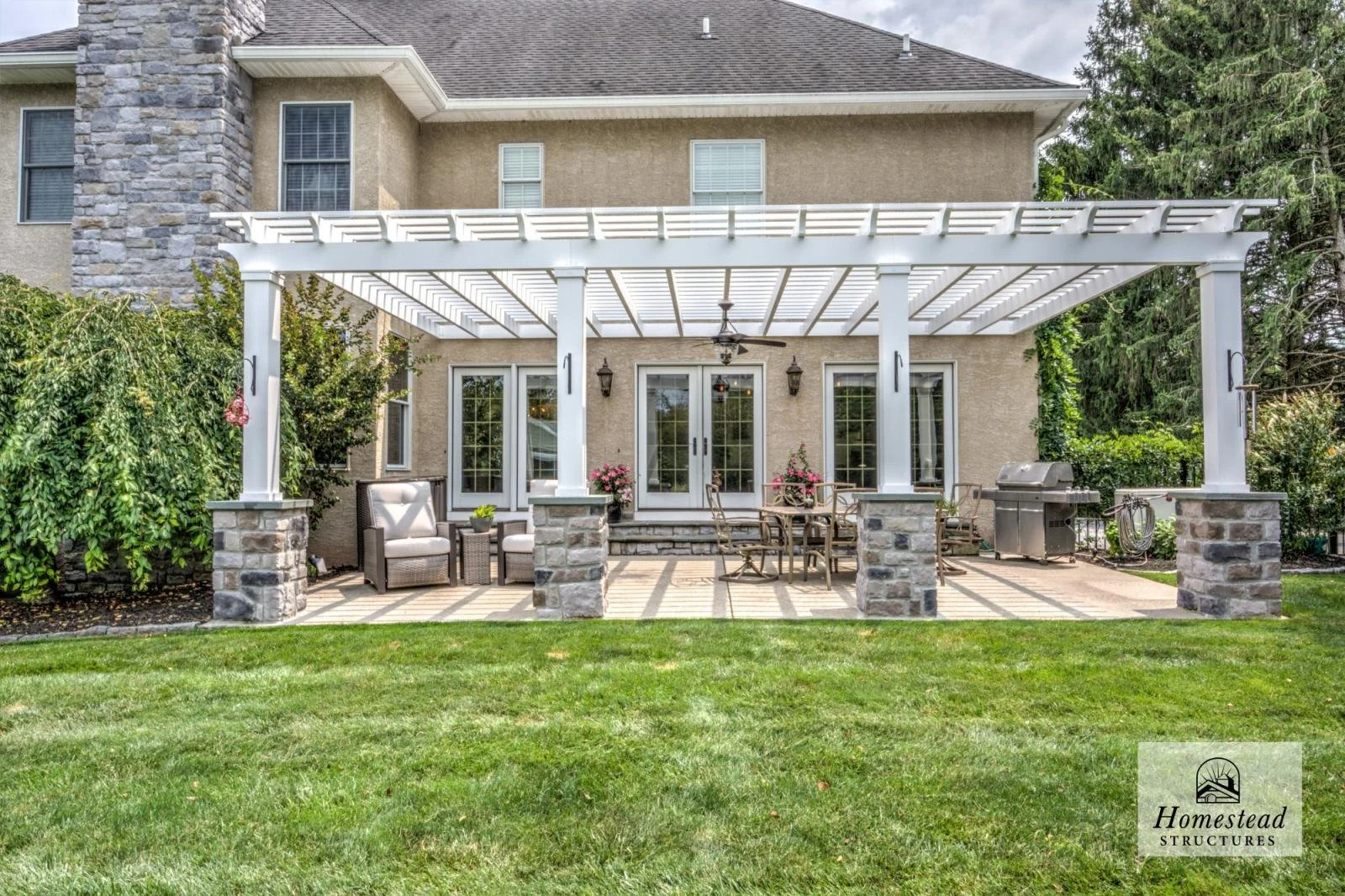 A backyard patio with a white pergola, outdoor furniture including chairs, a table, and a grill, with a house in the background. The area is landscaped with grass and shrubs.