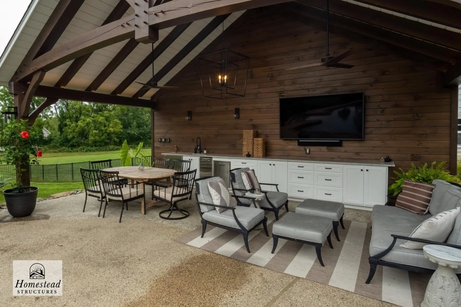 Covered outdoor living space with seating area, dining table, television, and kitchen counter, featuring wood ceiling and wall, greenery in background.