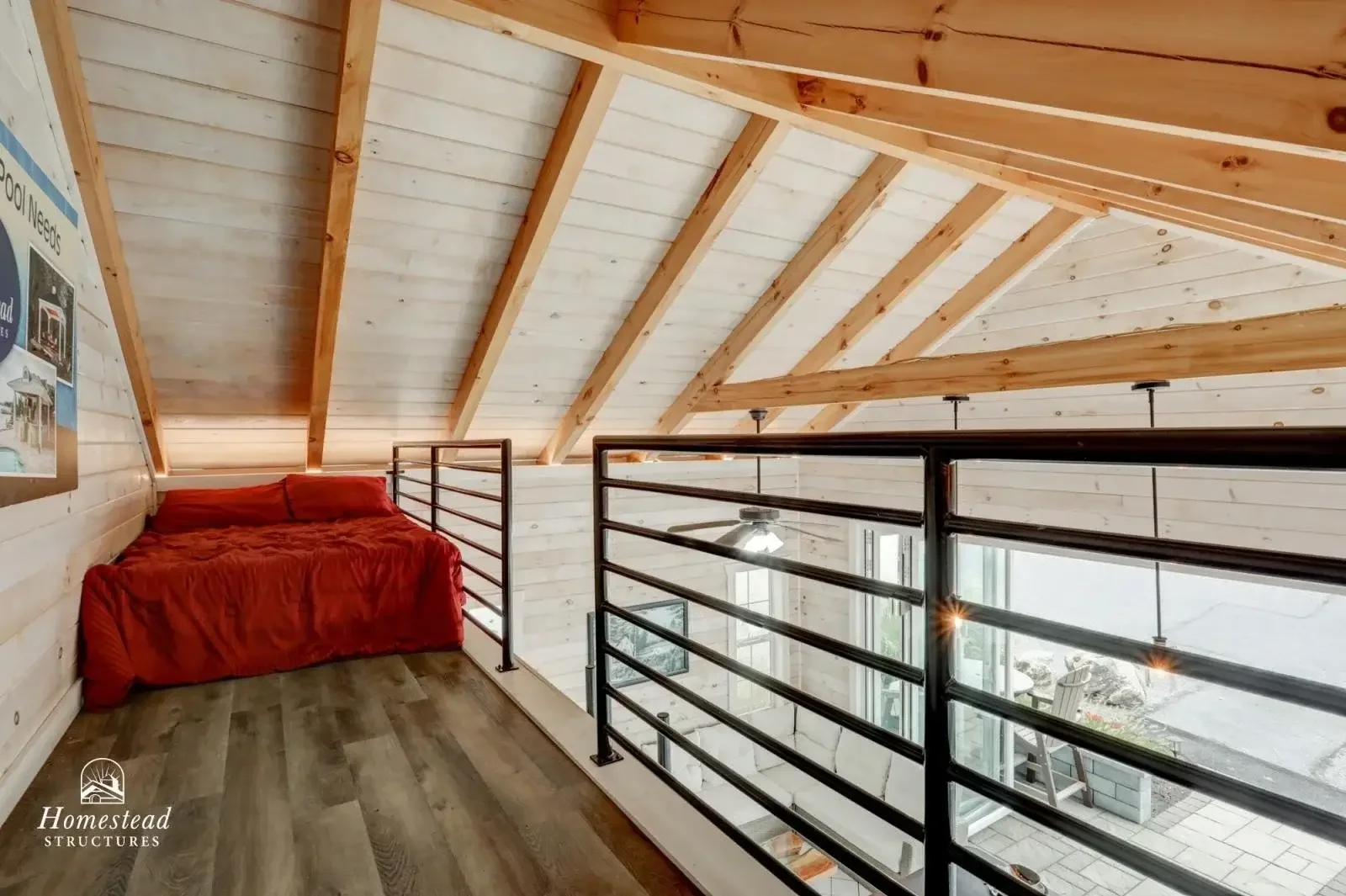 View from inside a kitchen and living area looking out to a patio and garden. The room features a vaulted wooden ceiling, a ceiling fan, a white L-shaped sofa, and a wooden and blue kitchen island set for dining. The patio outside has outdoor seating