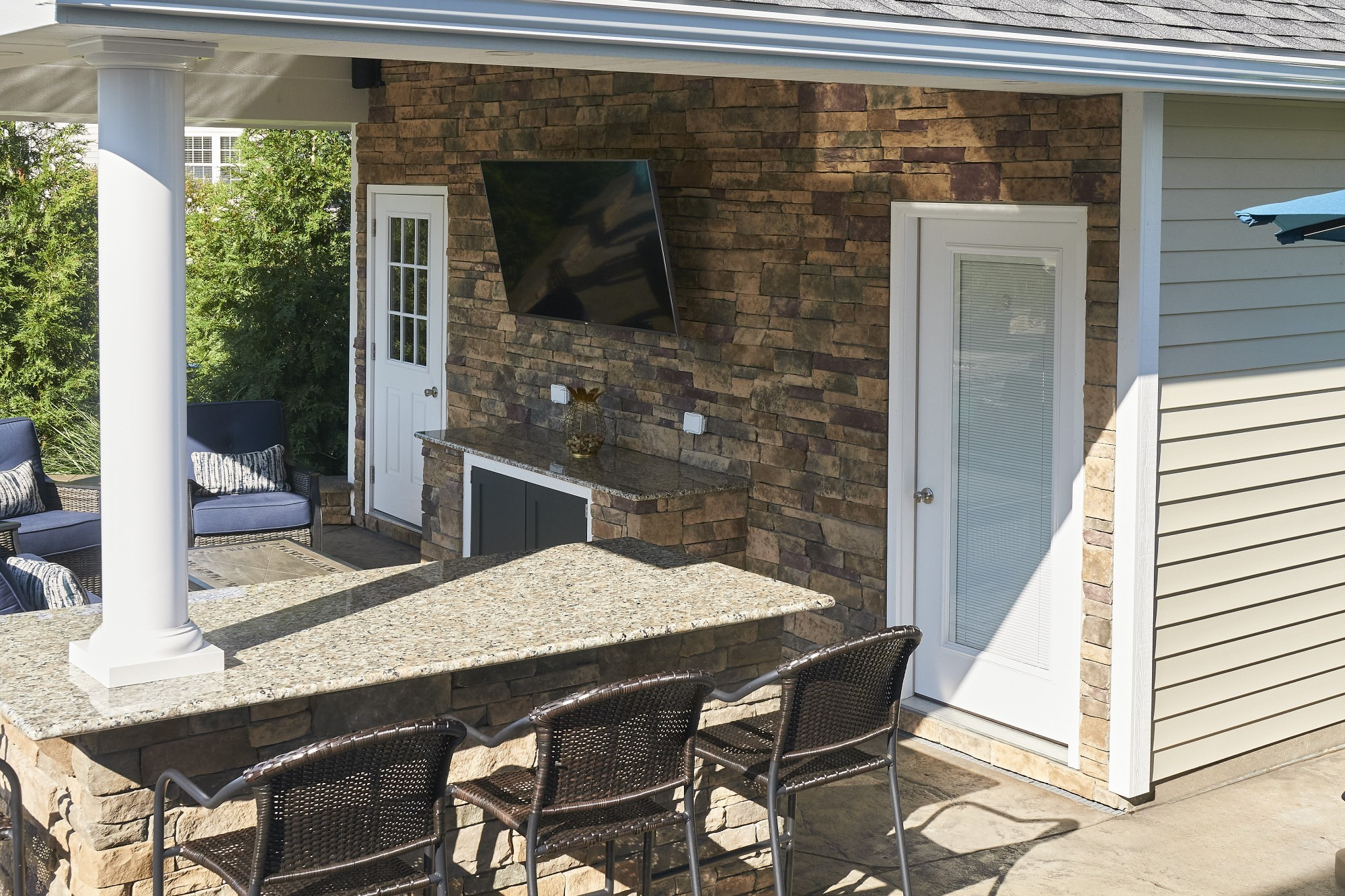 Outdoor patio with wicker chairs, a stone counter, a wall-mounted TV, and a brick wall with two white doors.