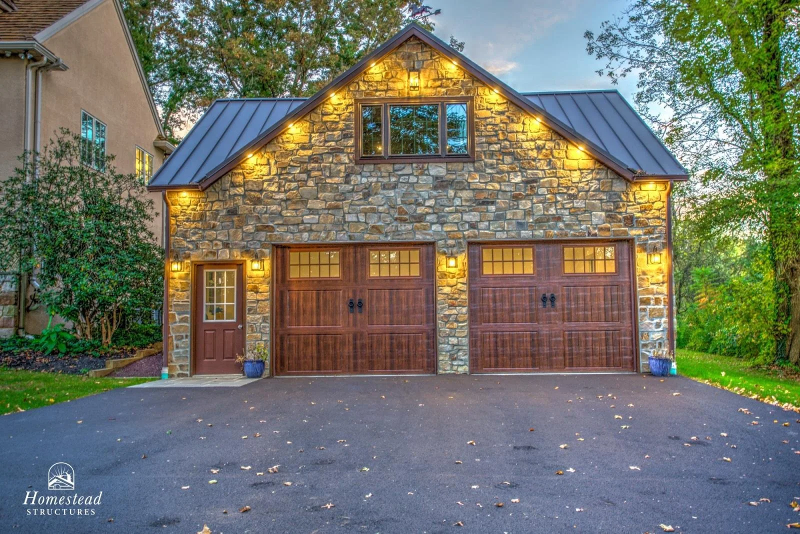 A two-story house with a stone exterior, wooden garage doors, and warm outdoor lighting, surrounded by trees.
