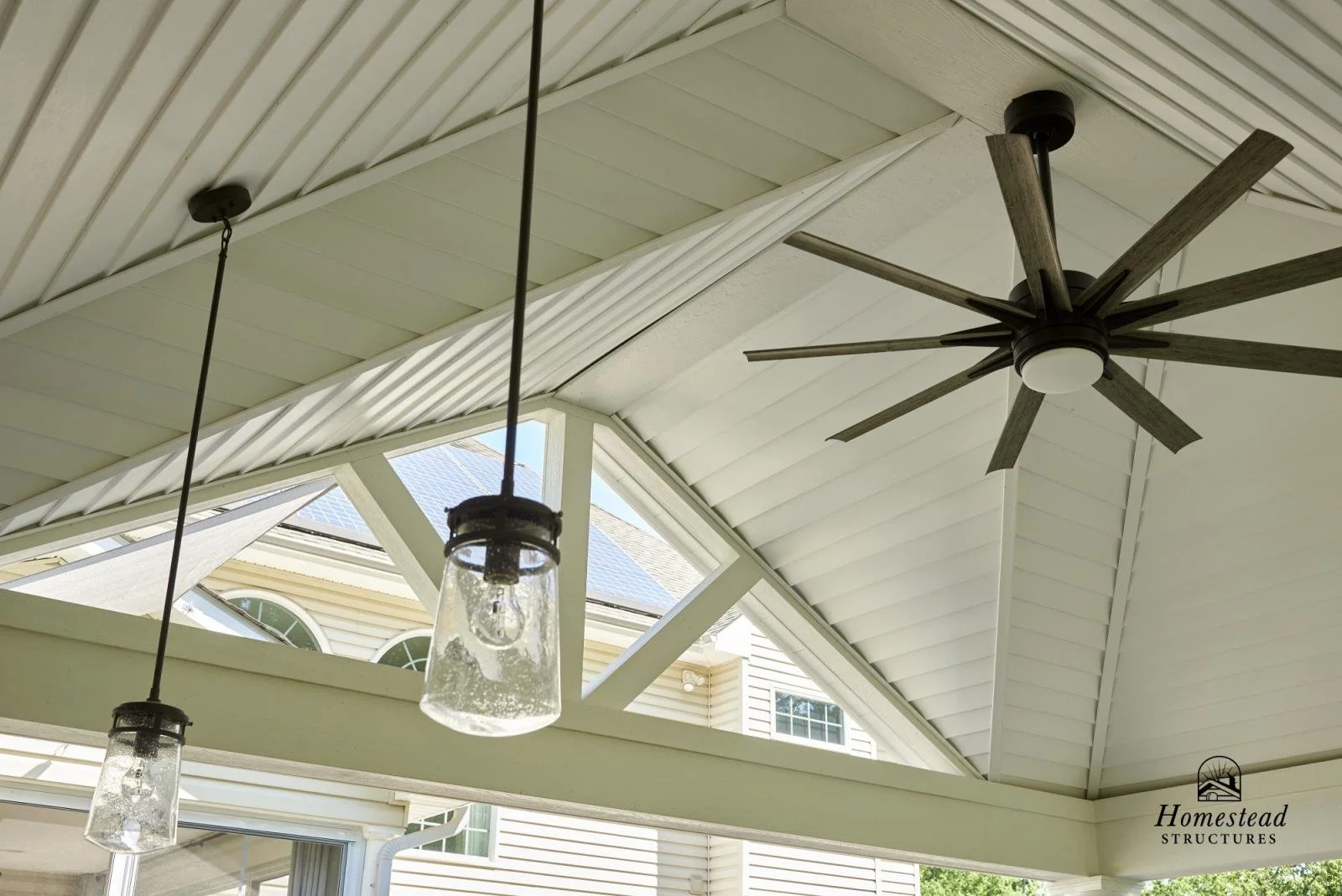 The ceiling of a covered porch featuring a large black ceiling fan, two pendant lights with glass shades, and a view of a house with solar panels on the roof.