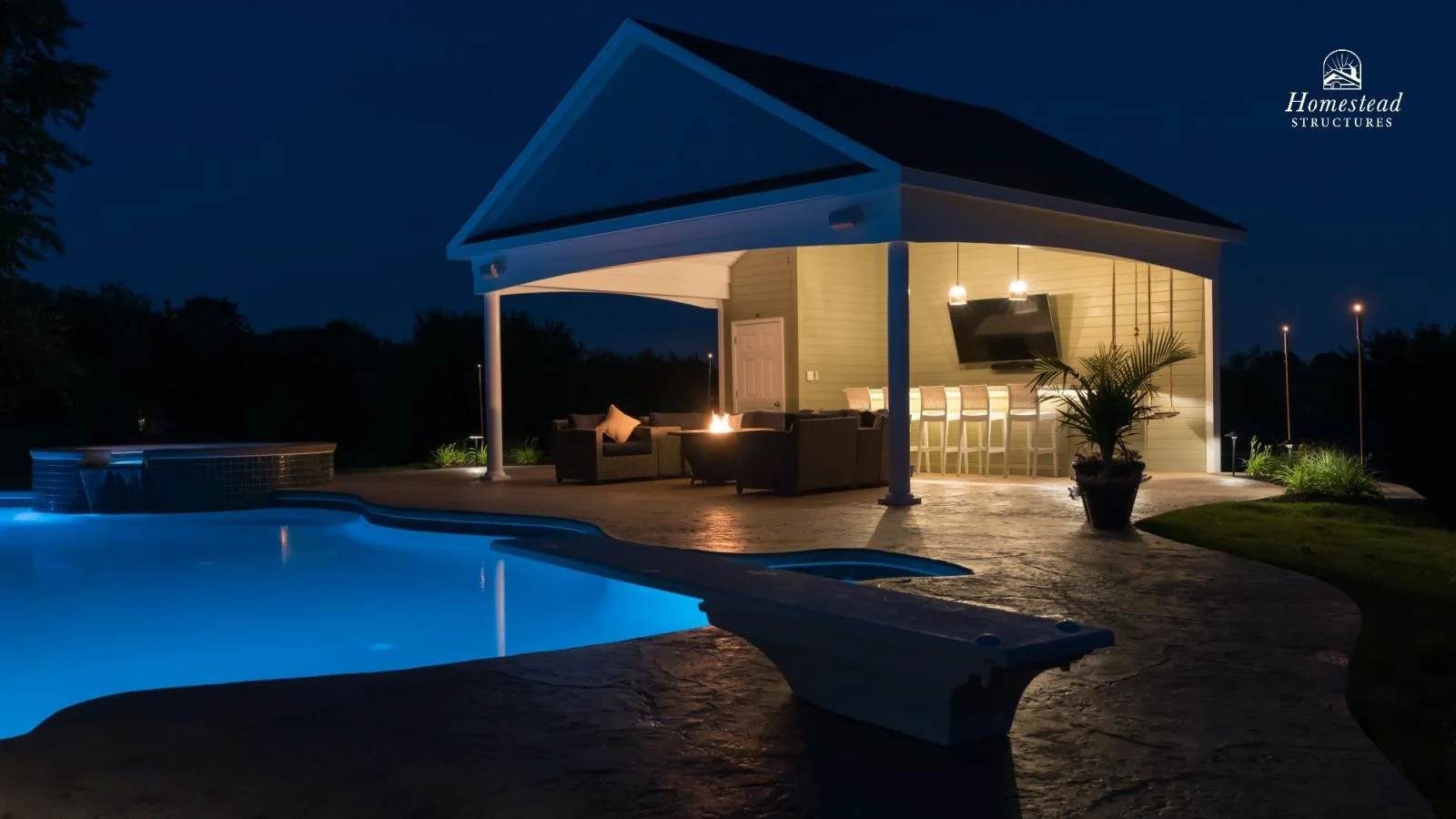 Night view of a lit outdoor pool area with a covered patio, seating, and bar area, with trees and dark sky in the background.
