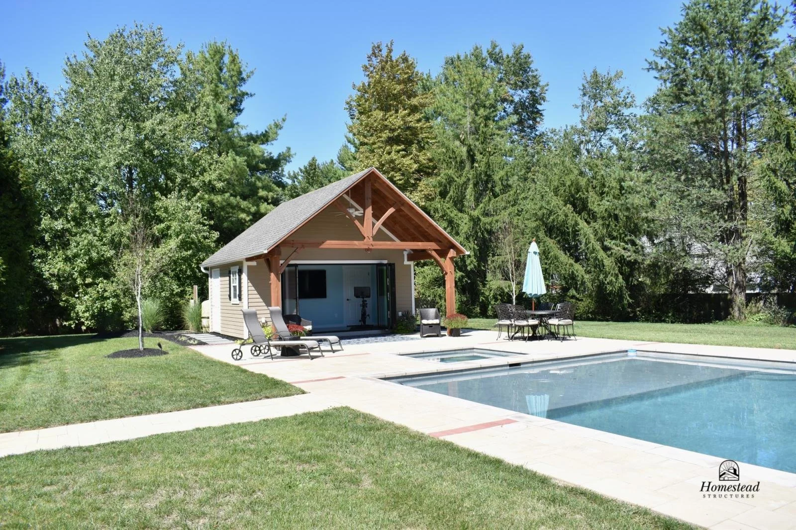 Backyard with swimming pool, patio with lounge chairs and table with umbrella, small house or pool house with open sliding doors, surrounded by green trees and grass.