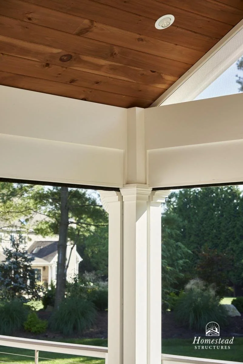 Close-up of a porch corner showing white columns, brown wooden ceiling, and large windows overlooking a garden with trees and shrubs.