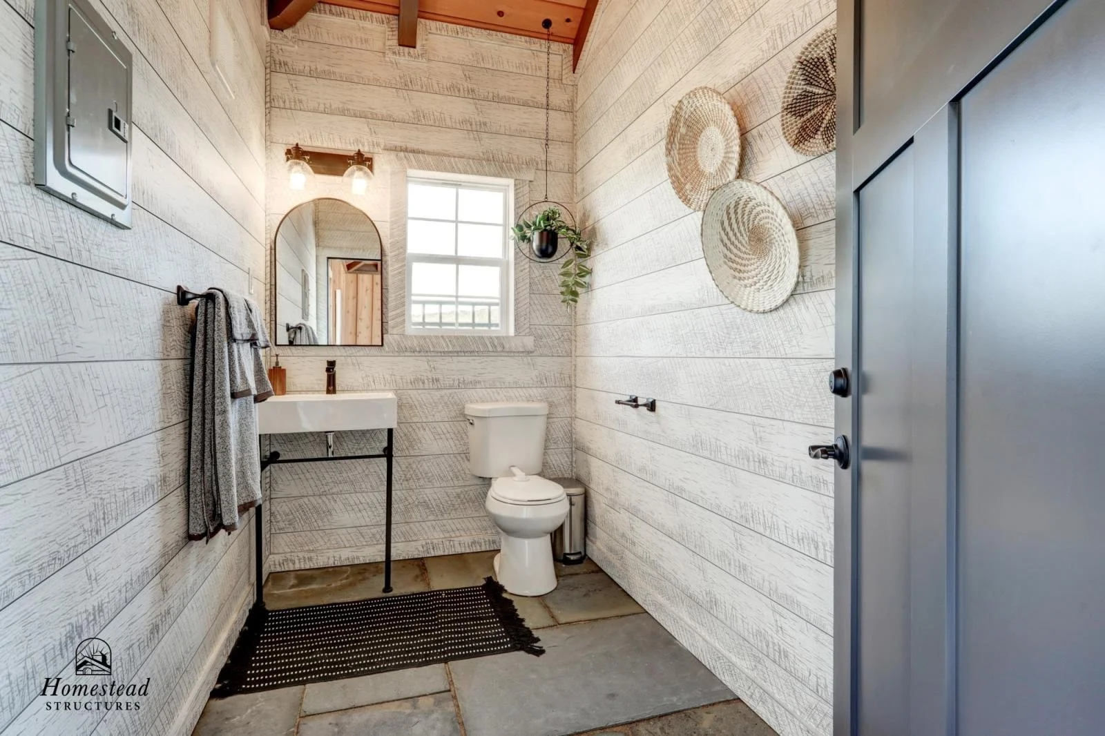Small rustic bathroom with whitewashed wooden walls, a small window, a white toilet, a black framed mirror above a white sink with black legs, a black towel rack, hanging woven baskets, a hanging plant, a black doormat, window light, and a gray metal