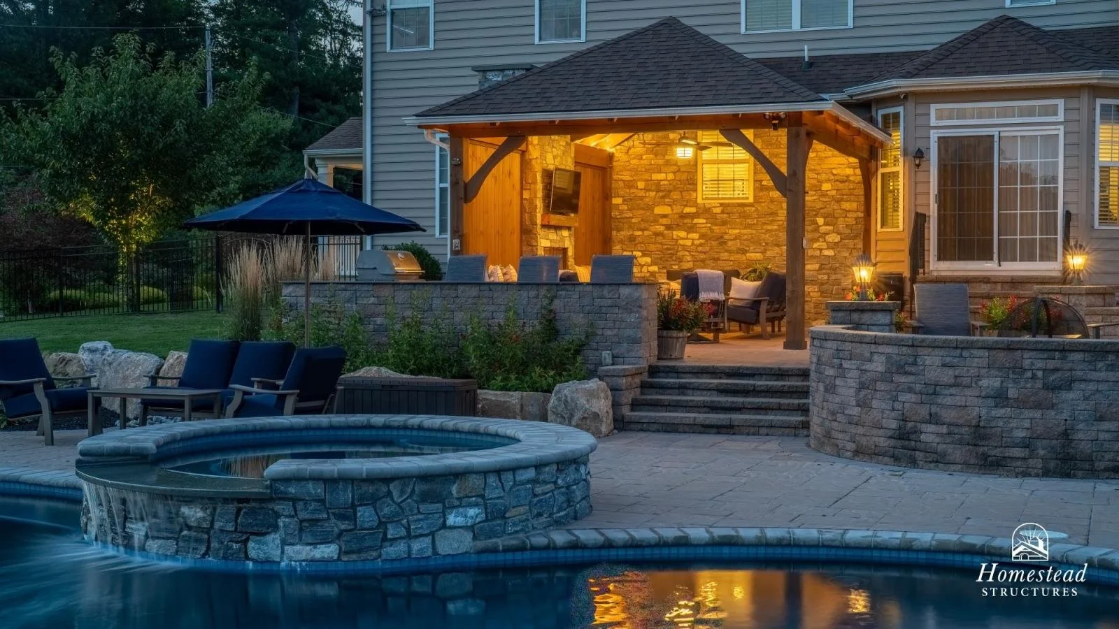 Backyard patio with outdoor seating, a hot tub, and a lit covered porch area at dusk.