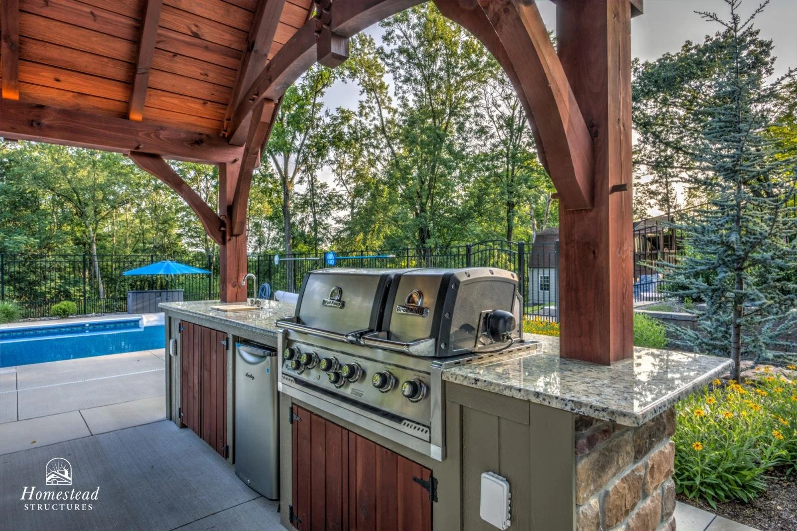 Outdoor kitchen with a grill and granite countertop, covered by a wooden pergola, overlooking a backyard with a pool and surrounded by trees.