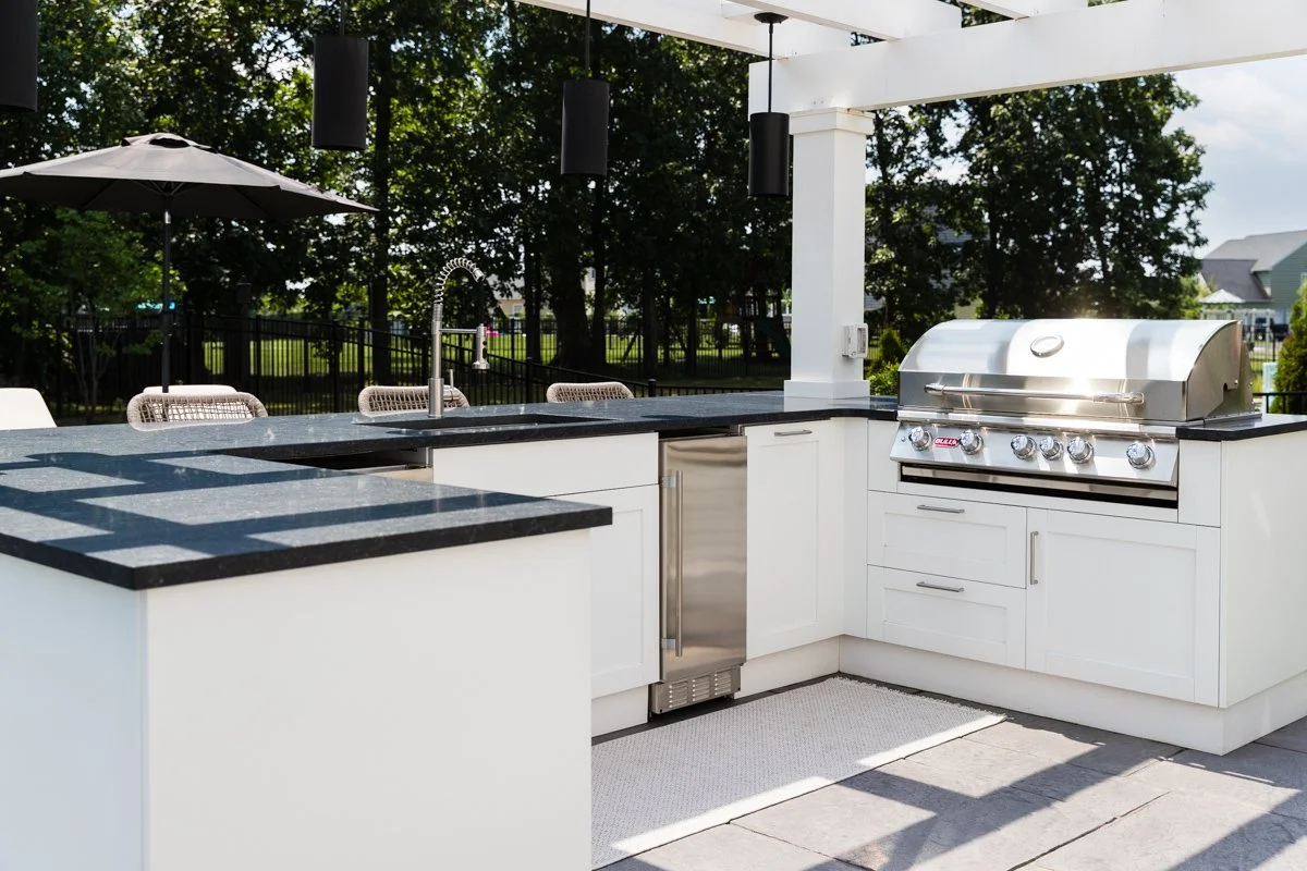 Outdoor kitchen with a stainless steel grill, white cabinets, a black countertop, and a sink, with seating and an umbrella in a backyard setting.