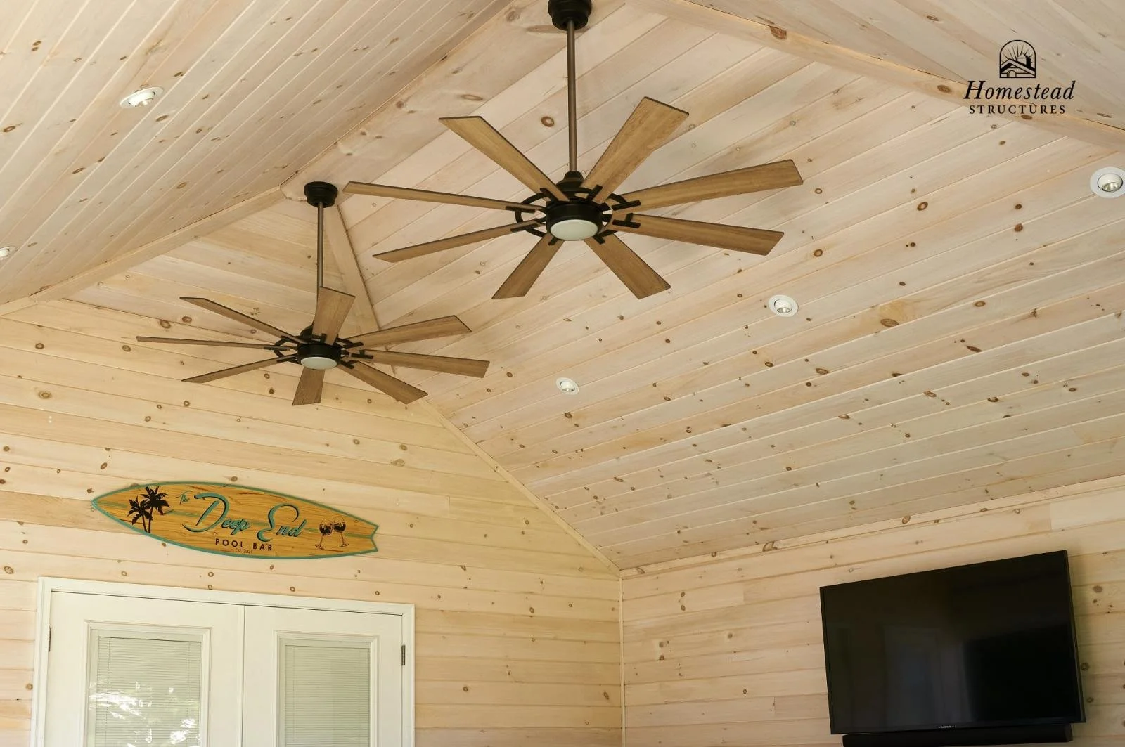 Wood-paneled ceiling with two ceiling fans, recessed lighting, a sign reading 'Deep End Pool Bar,' a door, and a mounted flat-screen TV.