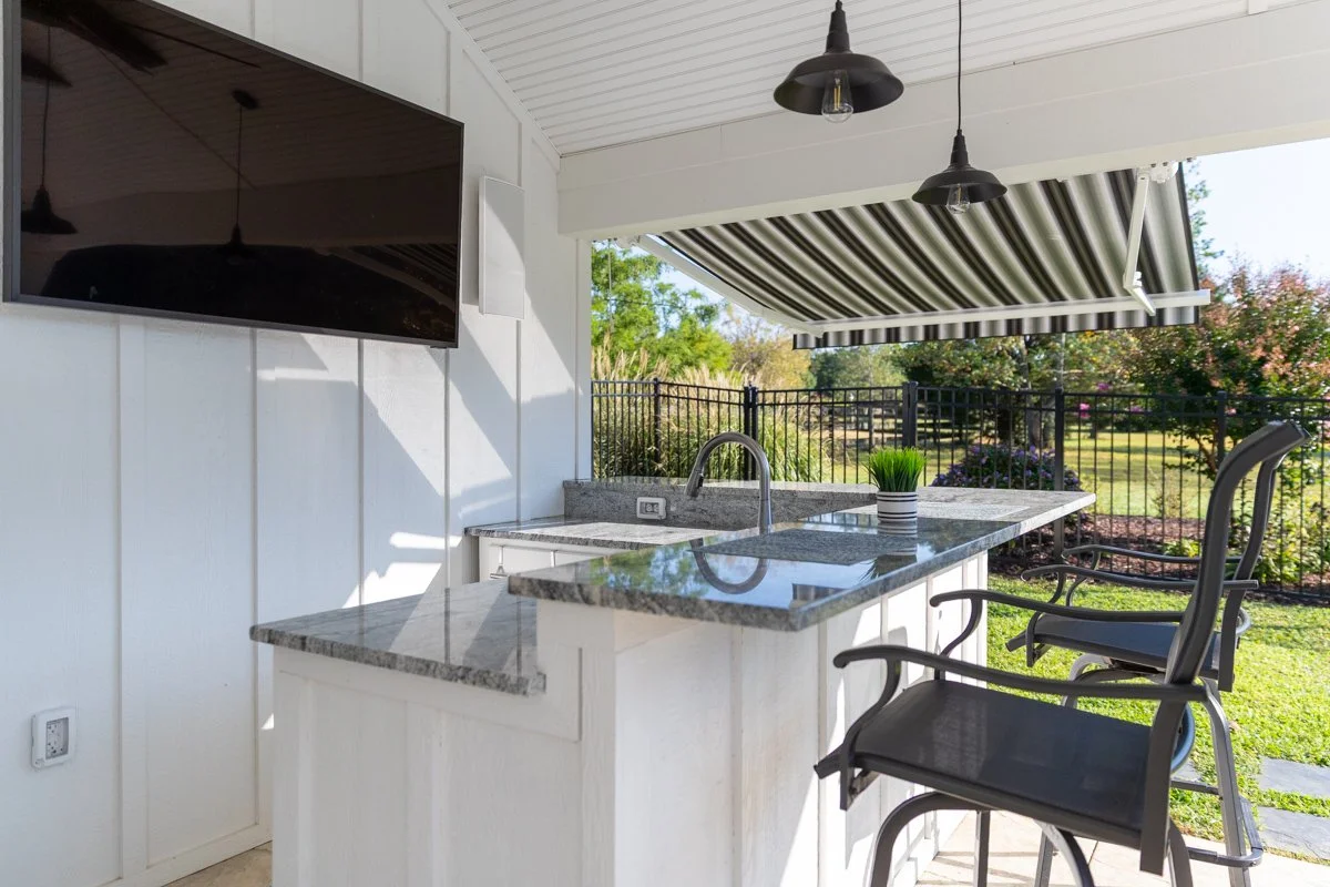 Outdoor kitchen with a granite countertop, sink, flat-screen TV on the wall, striped retractable awning, black metal chairs, hanging black pendant lights, and a view of a fenced backyard with trees and flowering shrubs.