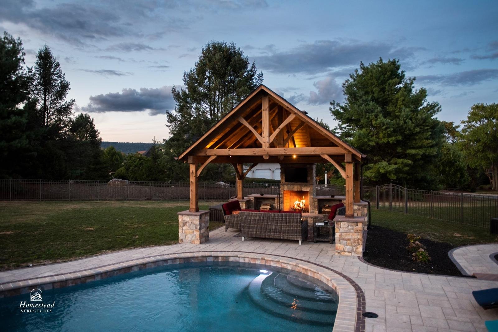 A backyard with a swimming pool and a wooden pavilion with a fireplace. The scene is during dusk with trees and a fence in the background.