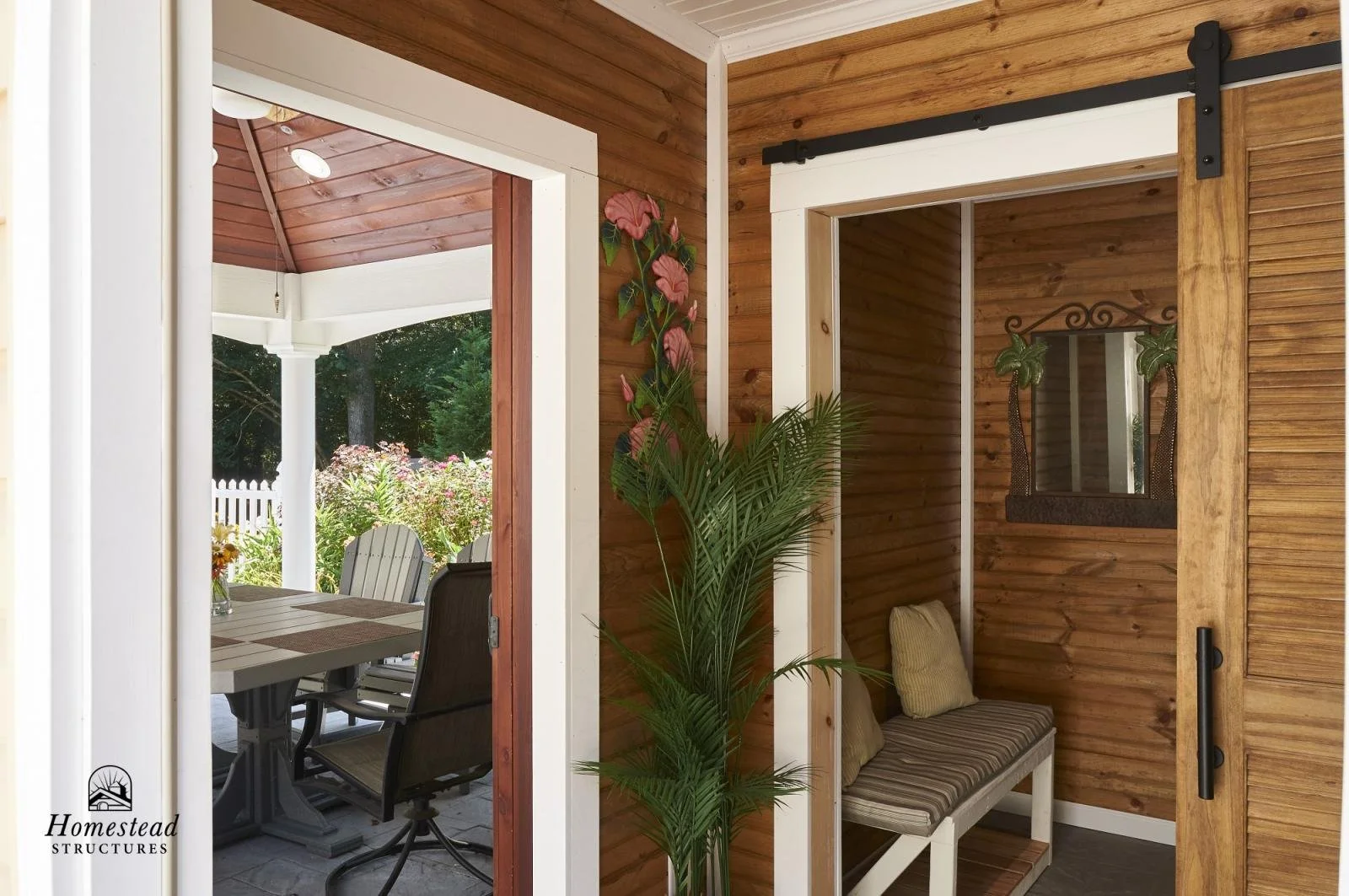 View of a porch with outdoor dining table and chairs seen through a sliding door, interior corner with wooden walls, a plant, and a window with a decorative mirror.
