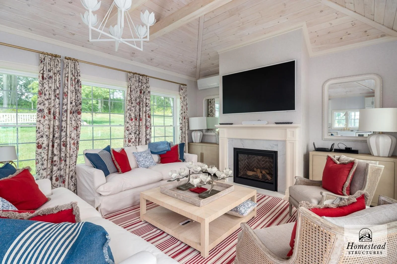 Living room with white sofas, red and blue pillows, striped rug, a wooden coffee table with a decorative centerpiece, large windows with floral curtains, a fireplace with a TV mounted above, and a mirror and lamp on side tables.