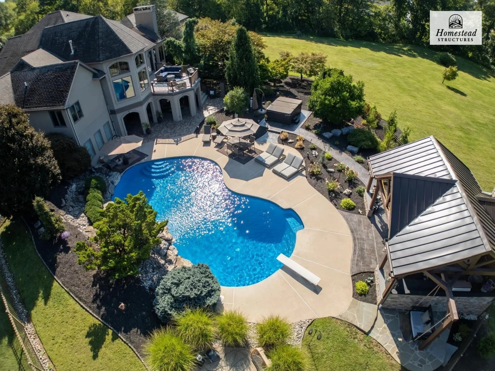 Aerial view of a backyard with a swimming pool, lounge chairs, a hot tub, a covered patio area, landscaped gardens, and a large lawn adjacent to a multi-story house with balconies.