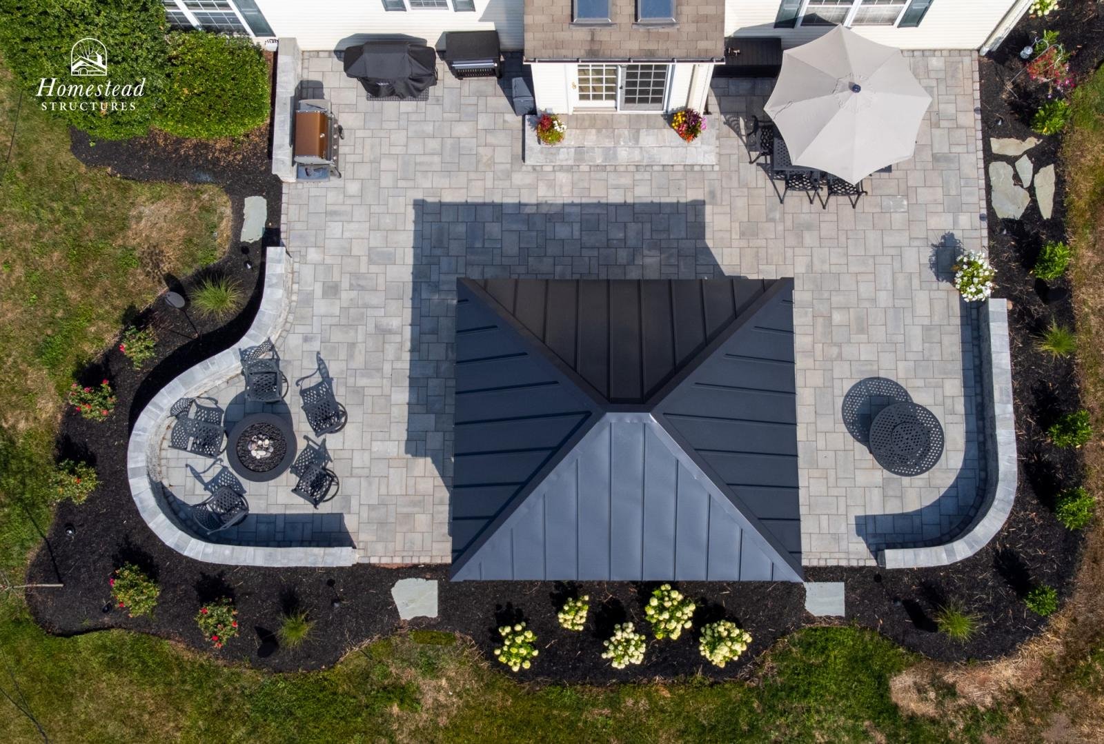 Aerial view of a backyard patio with outdoor furniture, a fire pit, an umbrella, and surrounding garden beds with plants and flowers.