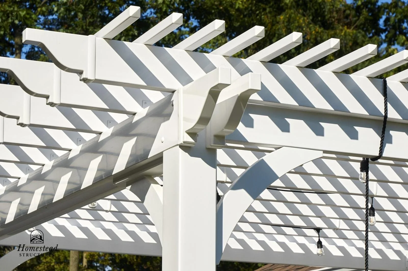 Close-up of a white wooden pergola with string lights hanging, casting shadows on the structure, against a background of trees and blue sky.