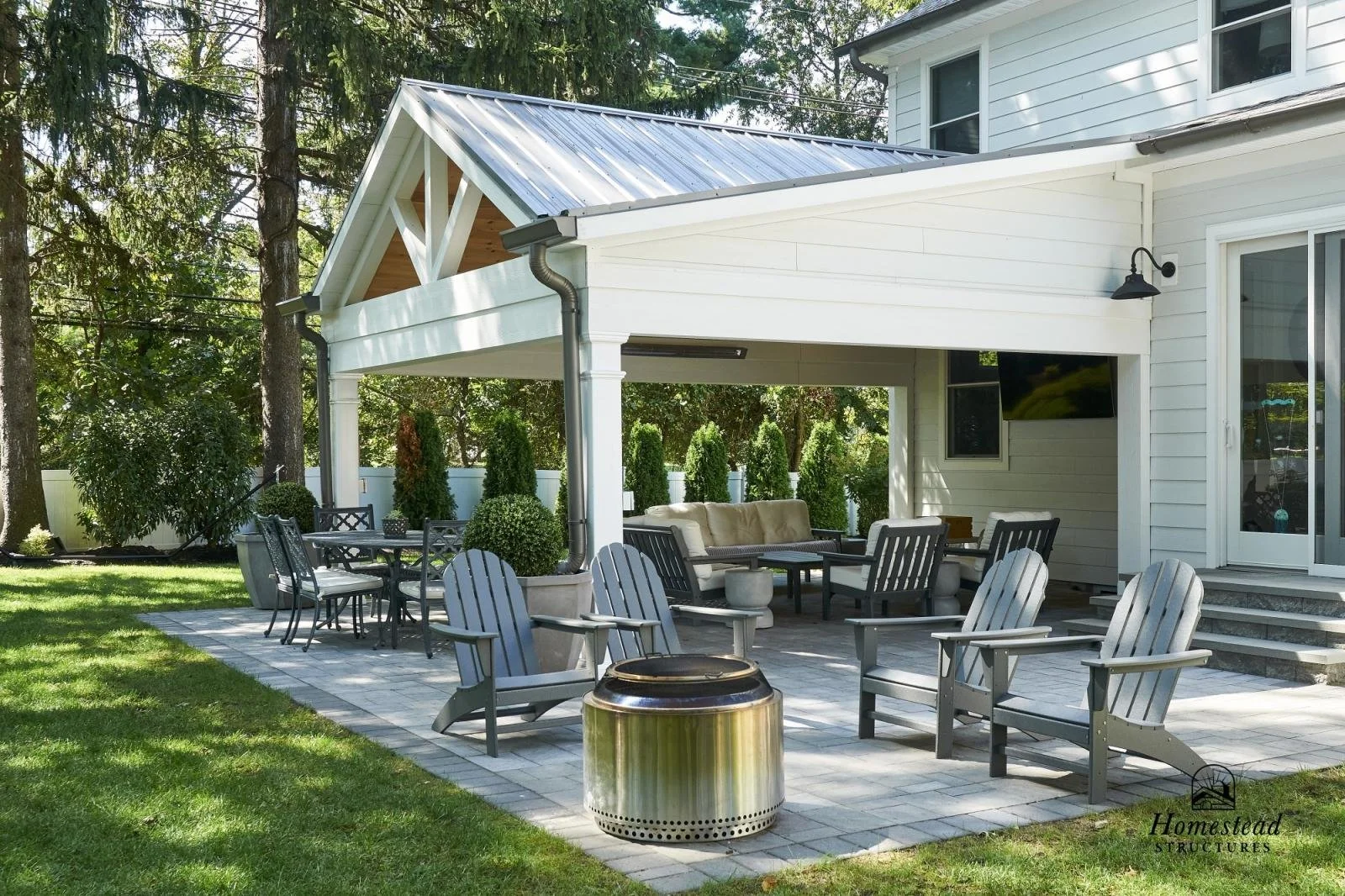 Back patio of a house with outdoor seating, including chairs, a sofa, a table, potted plants, and a fire pit, with a green lawn and trees in the background.