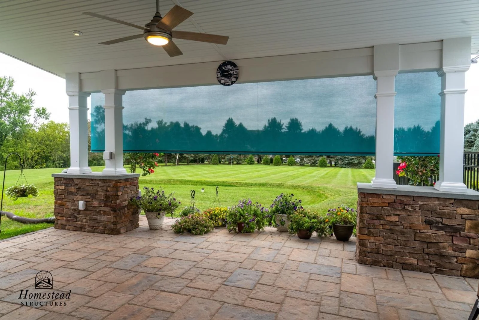 View of a covered porch with brick columns and a ceiling fan, overlooking a spacious, well-manicured lawn with flowers in pots, trees, and a blue shade screen.