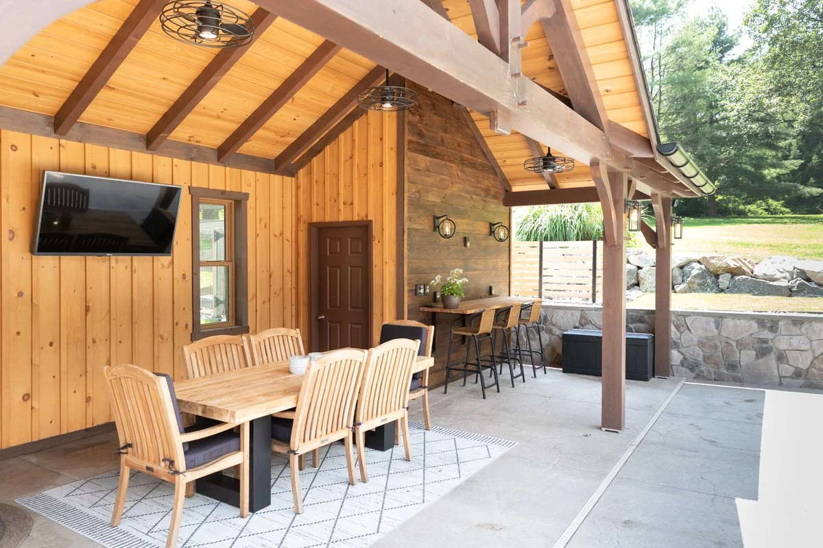 Outdoor patio with wooden walls and ceiling, featuring a dining table with eight chairs, a bar area with three chairs, wall-mounted lights, and a flat-screen TV, with a view of a grassy yard and rocks in the background.