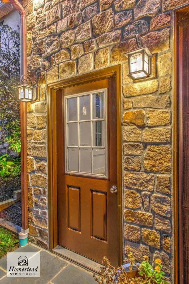 Stone wall exterior with a brown door and two wall-mounted lanterns illuminating the entryway.