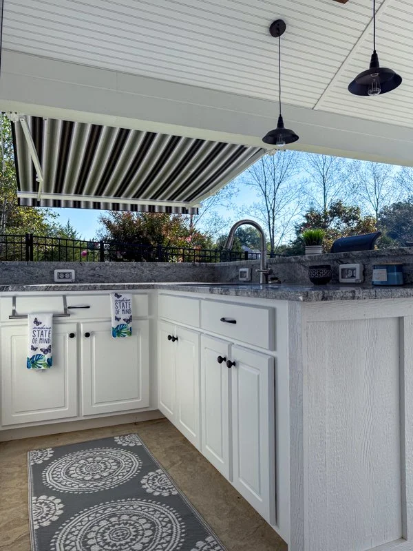 Outdoor kitchen with white cabinets, granite countertop, sink, and a retractable striped awning. There are hanging black pendant lights and a gray decorative rug on the floor. The background shows trees and a fence.