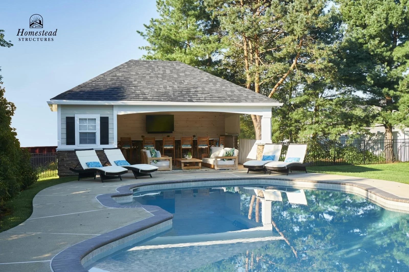 Backyard with a swimming pool, and a covered patio area with lounge chairs, a dining table, and a TV. The patio is adjacent to a house with white siding and black shutters, surrounded by green trees and grass.
