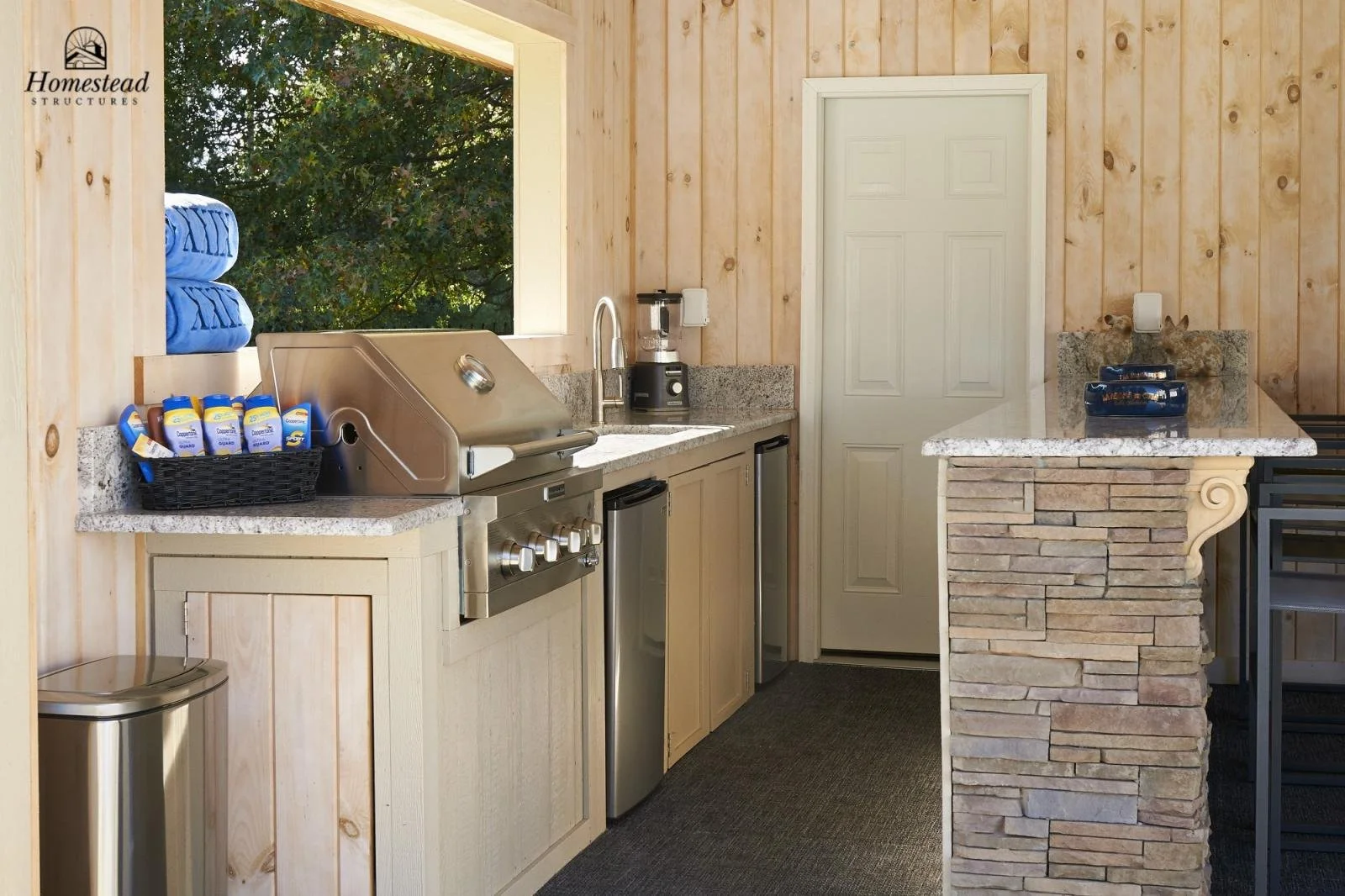 Outdoor kitchen with a granite countertop, a built-in stainless steel grill, a blender, and a small refrigerator, featuring light wooden paneled walls, a large window showing trees outside, and a stone bar with a decorative volute detail. Blue towels