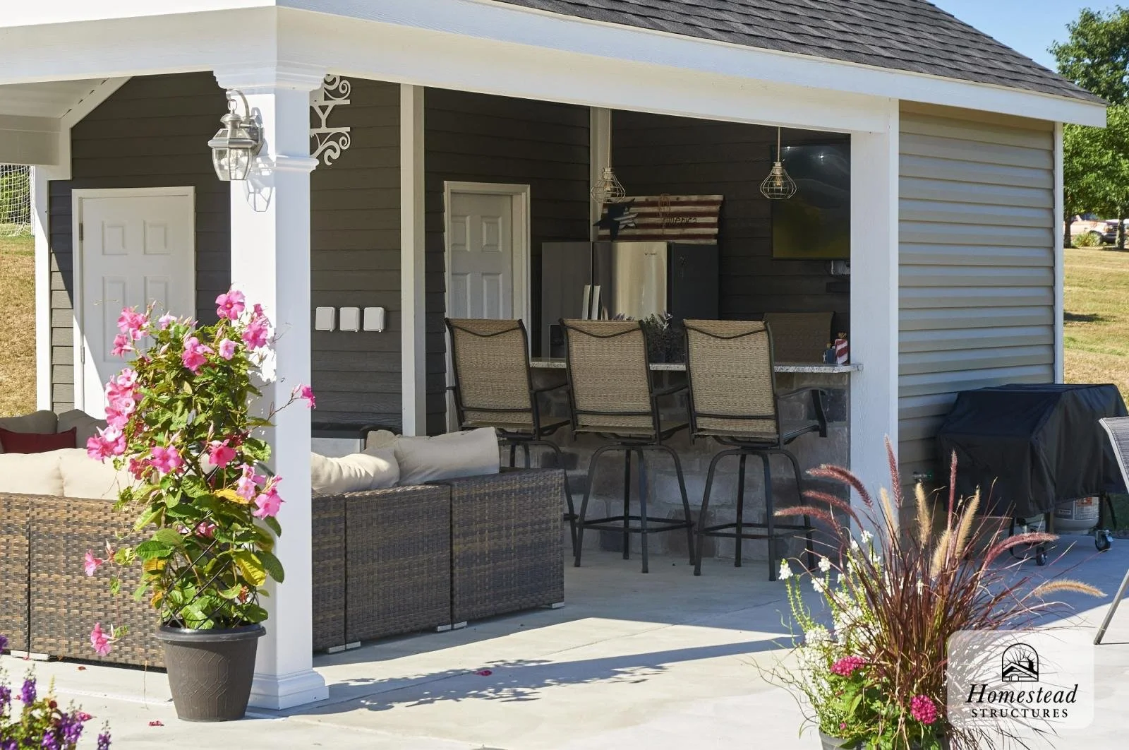 A covered outdoor patio with bar stools, a seating area with wicker furniture, potted pink flowers, and a black barbecue grill, attached to a gray house with white trim.